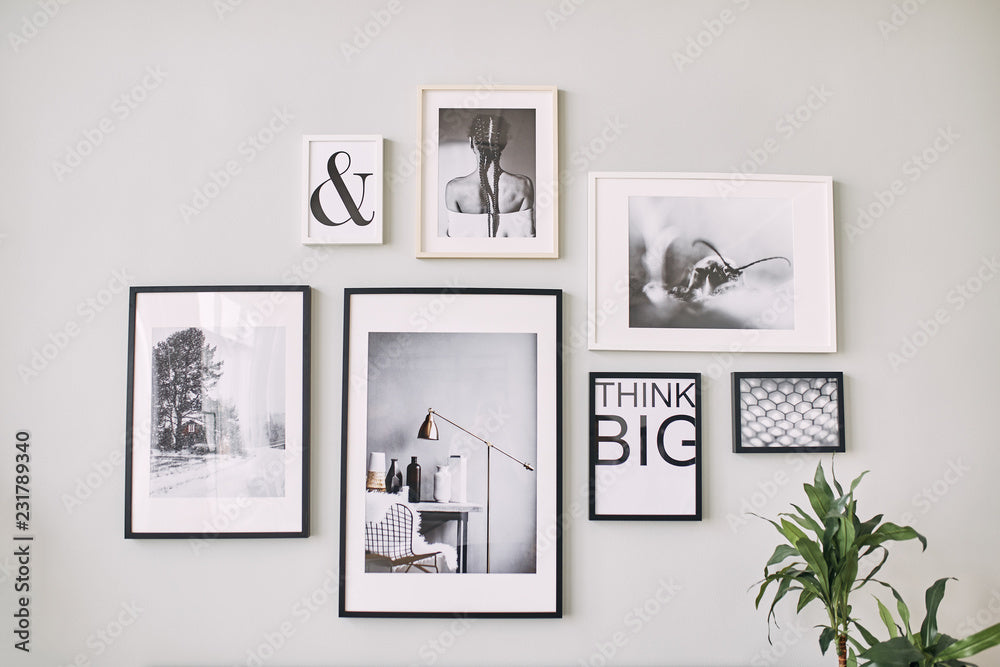 Photo of a white wall with a poster in the center in a black frame, with a cluster of five photographs in black frames on either side