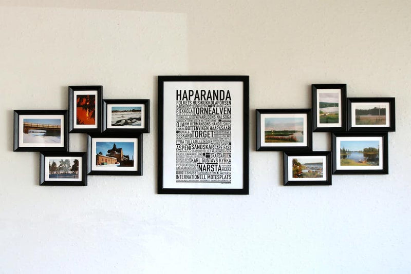 Photo of a white wall with a poster in the center in a black frame, with a cluster of five photographs in black frames on either side