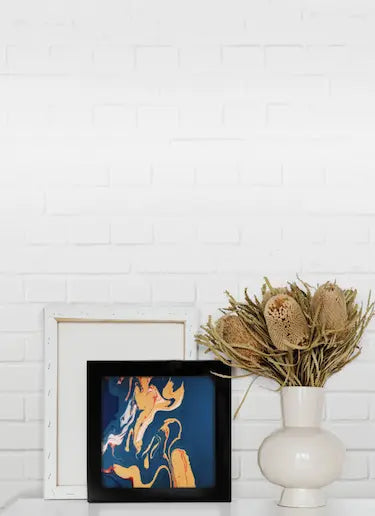 A black picture frame with a border displays an abstract picture on a table next to a vase of dried flowers.